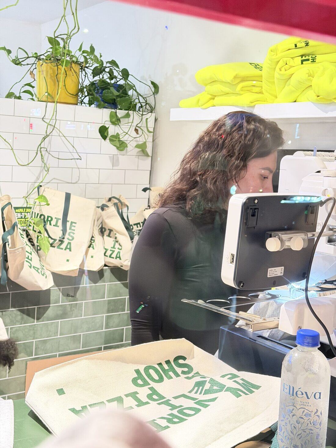 Merch Troop operator running an embroidery machine behind the counter of a brand popup with canvas totes hanging nearby