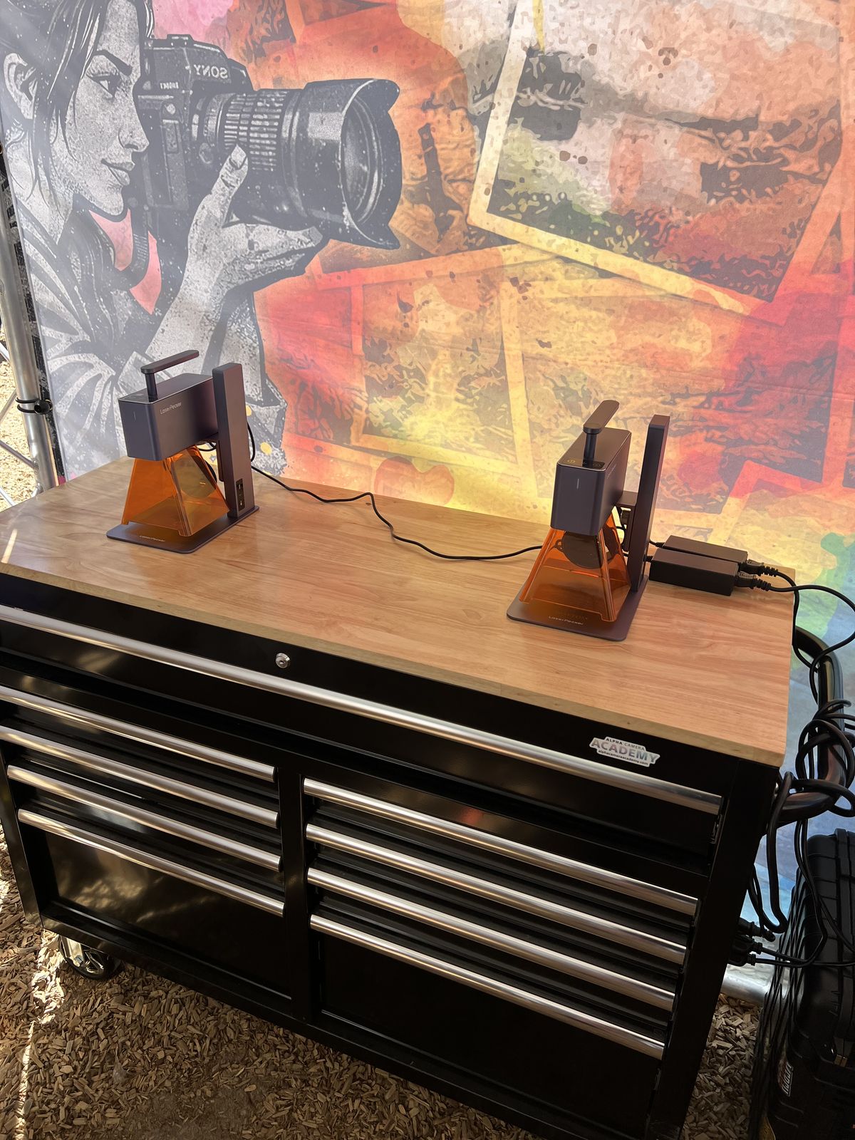 Two compact laser engravers set up on a wood-top work cart for personalizing goods at an outdoor festival